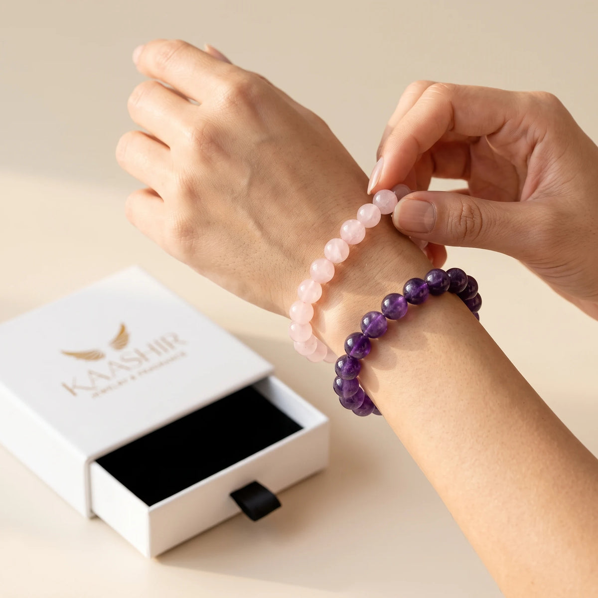 Woman putting on Kaashir rose quartz and amethyst bracelets beside an open gift box
